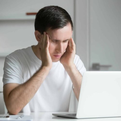 Young man, hands at his temples, at the white desk, laptop near Estresse e ansiedade afetam saúde bucal